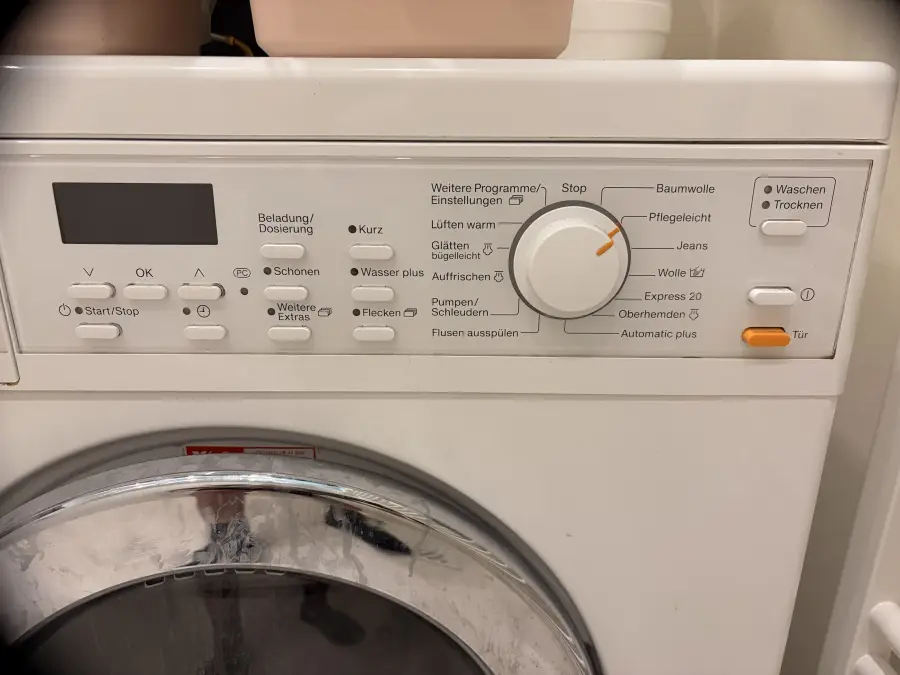 Das Bedienfeld: links Display und Start/Stop, Mitte der Drehknopf für das Programm, rechts Waschen/Trocknen, Ein/Aus und die orange Taste „Tür“.